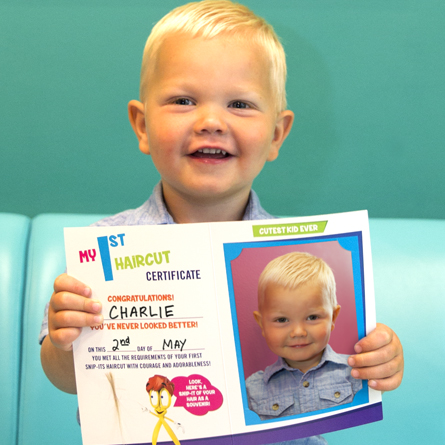 A young boy smiling and holding up his First Haircut Certificate from Snip-its.