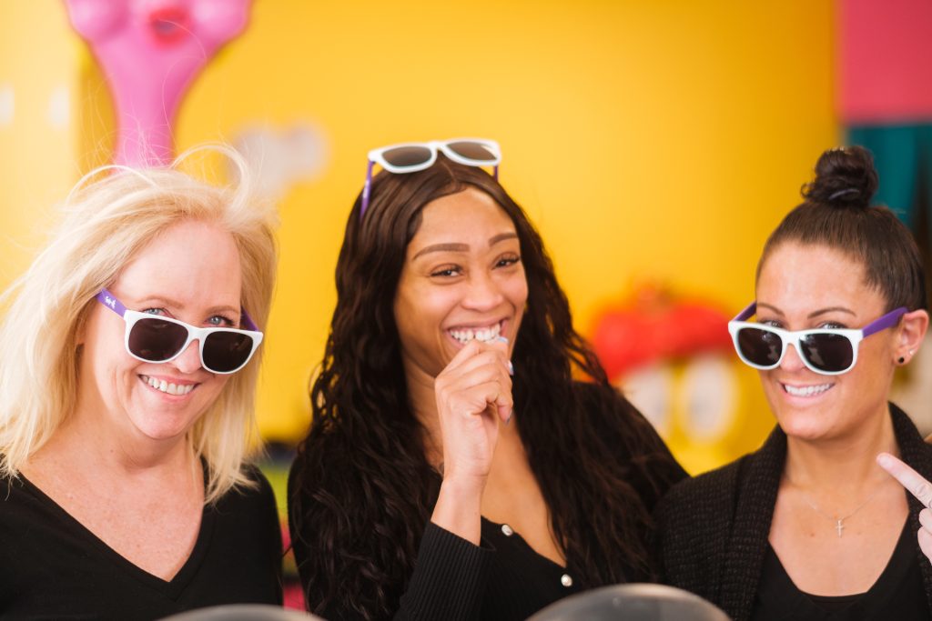 3 women with sunglasses standing in front of a yellow background laughing and smiling